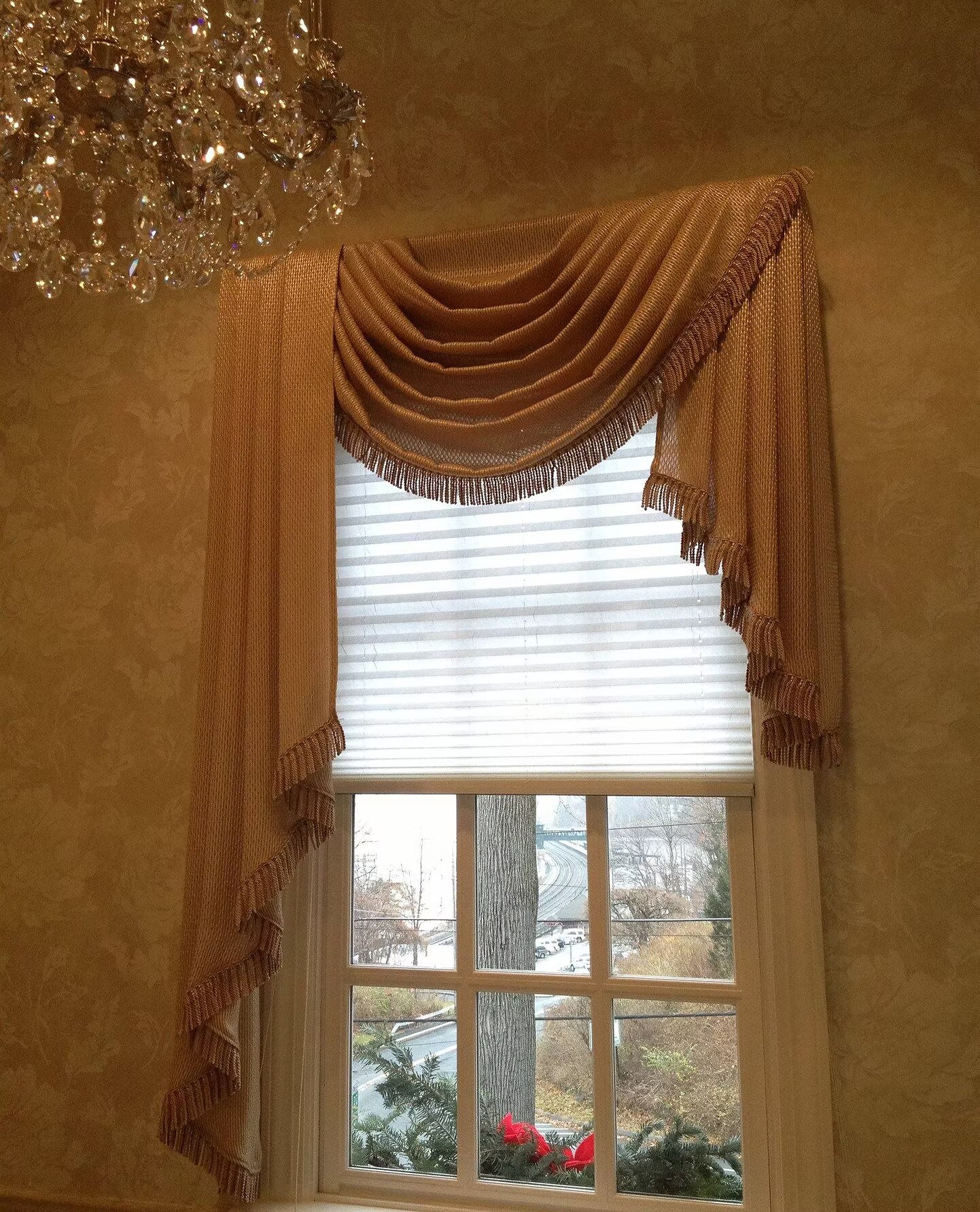 A custom gold swag valance with bullion fringe over a tall window, beneath a crystal chandelier